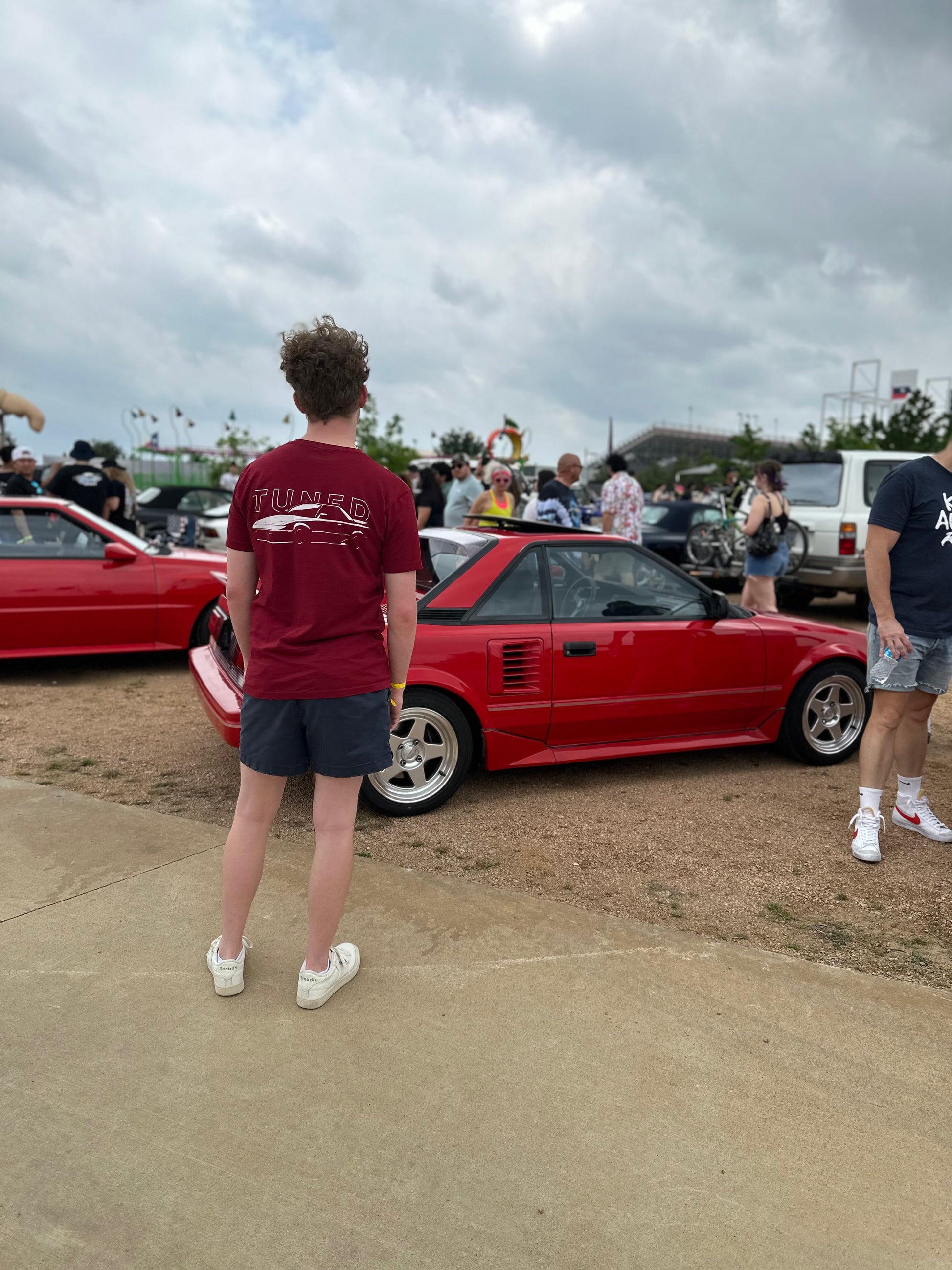 A model stands facing a red toyota Mr2 sports car with their back to the camera. They are wearing the mens red mr2 shirt.